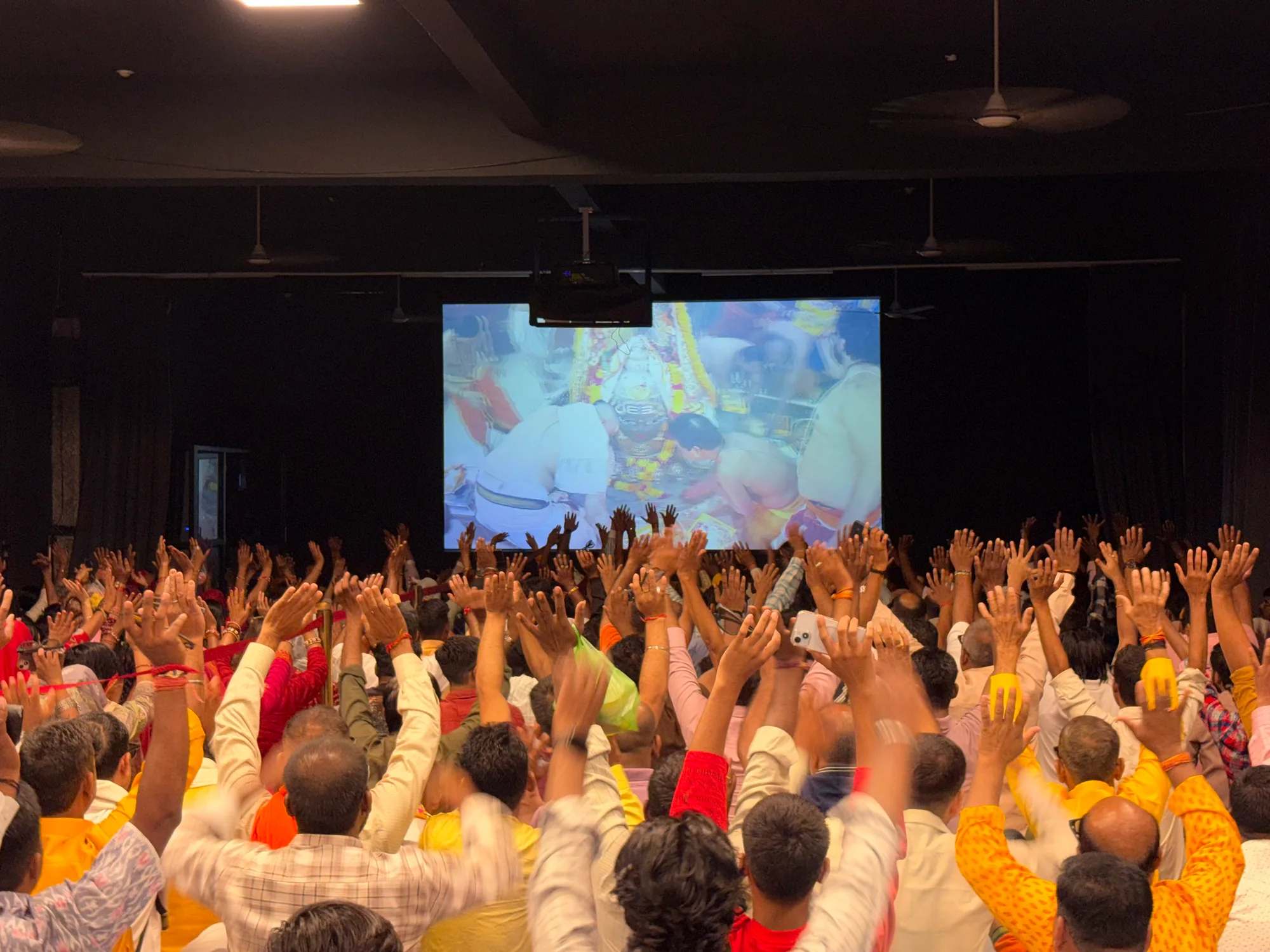 A hall of devotees with raised hands, the live Bhasma Aarti from the sanctum on the large screen